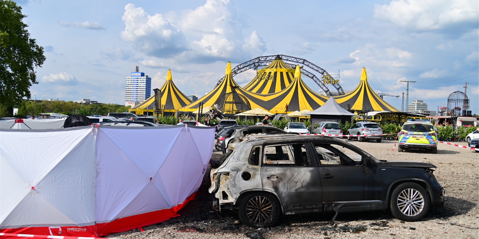 Ausgebrannte Fahrzeuge stehen nach dem Absturz eines Kleinflugzeugs an dem aufgestellten Sichtschutz, mit dem das Flugzeugwrack abgeschirmt wurde. Im Hintergrund steht das Zelt des zirkus Flic Flac.