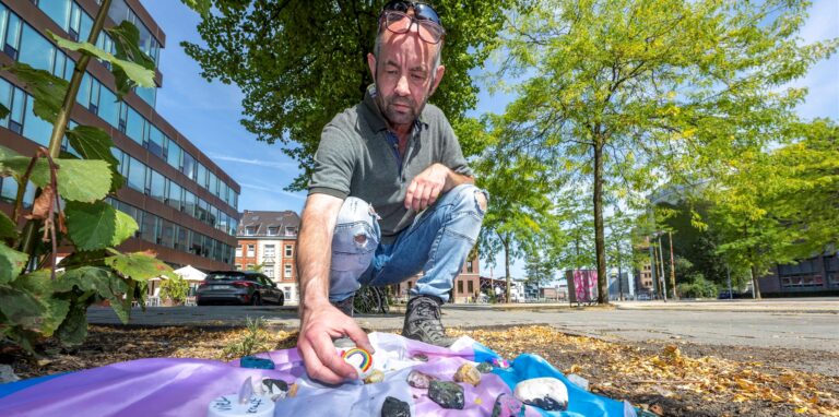 Felix Adrian Schäper vom Verein Trans*Inter*-Münster kniet vor einer Fahne mit Gedenksteinen, die Menschen auf dem Hafenplatz ausgelegt haben.