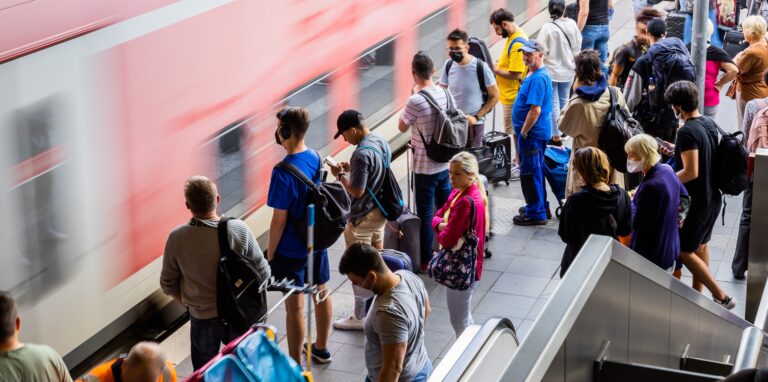 Menschen stehen am Bahnsteig, an dem gerade ein Zug vorbeifährt.
