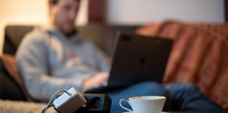 Ein Mann sitzt an seinem Laptop, im Vordergrund eine Kaffeetasse und ein Netzteil in einer Steckdose.
