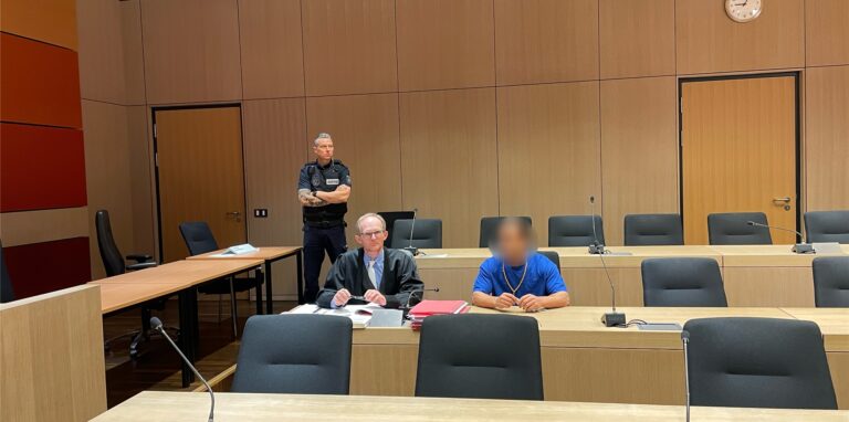 Drei Personen in einem Gerichtssaal. Linke ein stehender Justizbeamter, dann der Verteidiger und sein unkenntlich gemachter Mandant sitzend.