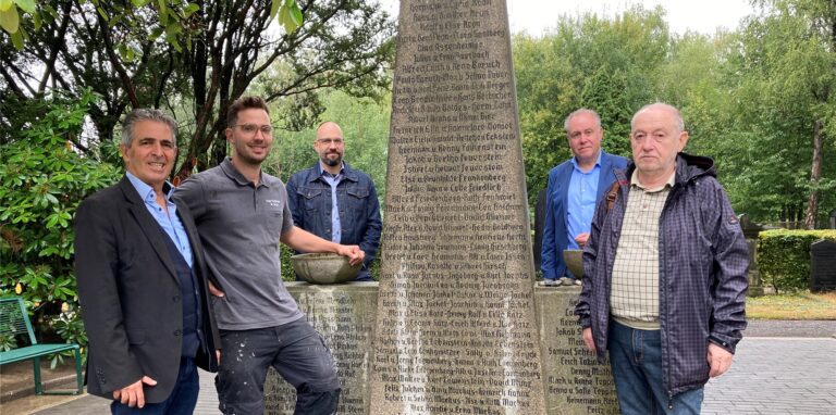 Fünf Männer stehen auf dem jüdischen Friedhof in Recklinghausen neben einem Mahnmal für Holocaust-Opfer.