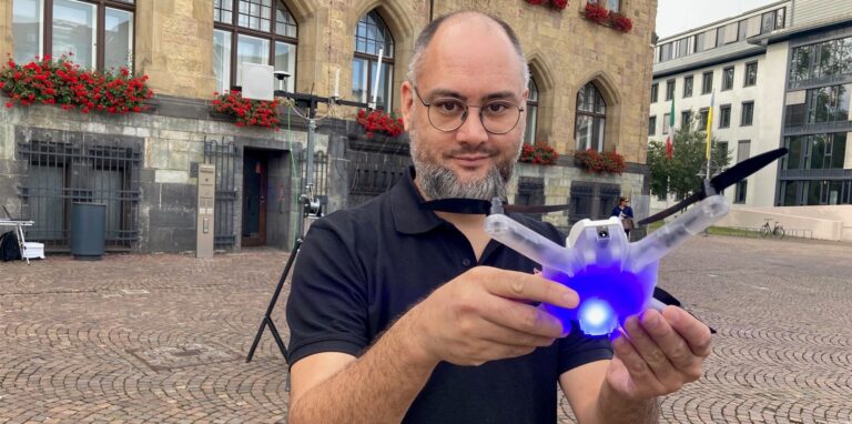 Ein Mann mit Bart und Brille hält eine blau leuchtende Flugdrohne in den Händen.