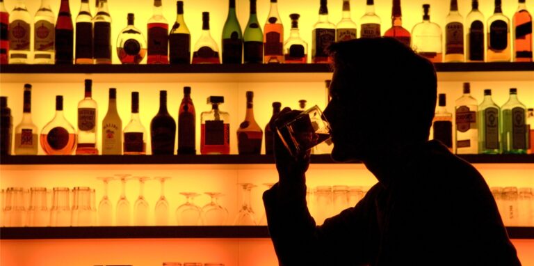 Ein als Schattenriss erkennbarer Mann sitzt in einer Bar vor einer beleuchteten Wand mit Flaschen und trinkt aus einem Tumbler-Whiskyglas.