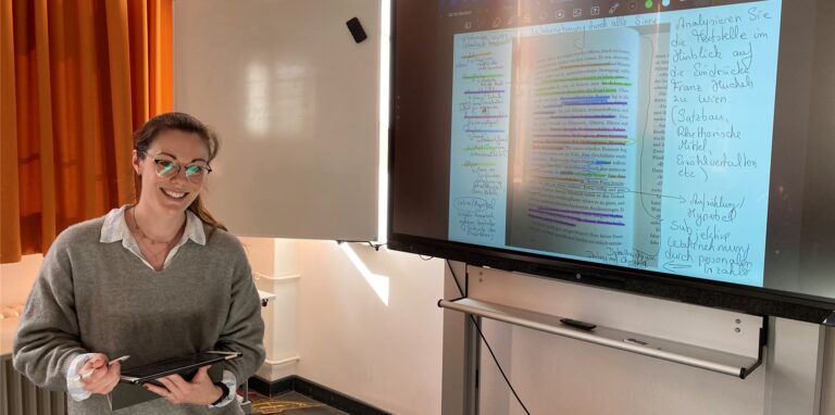 Lehrerin Sarah Weber mit einem iPad in der Hand vor einer digitalen Tafel.