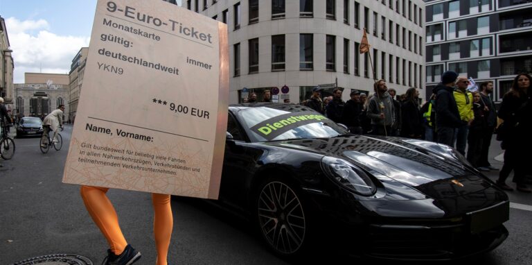 „Dienstwagen“ steht auf einem Porsche 911 während ein Demonstrant mit einem übergroßen „9-Euro-Ticket“ bei einer Protestaktion der Umweltschutz-Organisation Greenpeace vor der FDP-Bundeszentrale daneben steht.