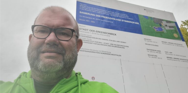 Reporter Jörg Müller vor einem Bauschild, auf dem zu lesen ist: Sanierung der Freibadanlage Stimbergpark.