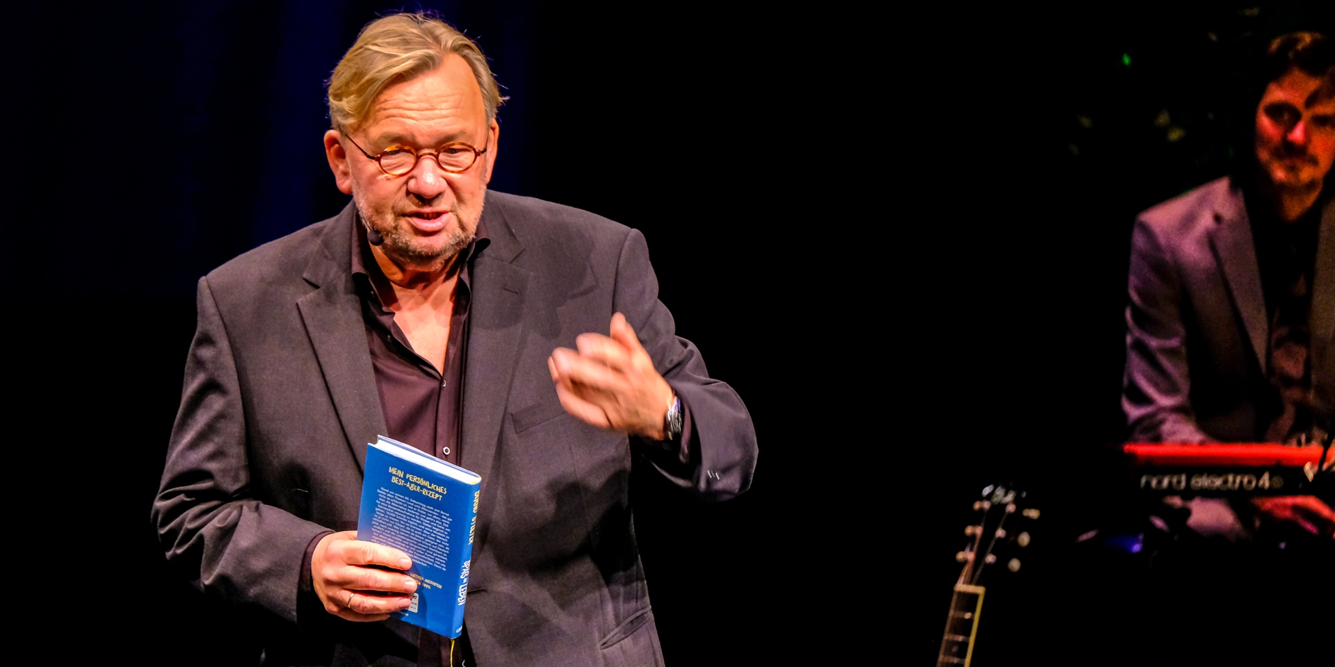 Bernd Stelter – mit Brille, Sakko und einem Buch in der Hand – steht auf einer Bühne.