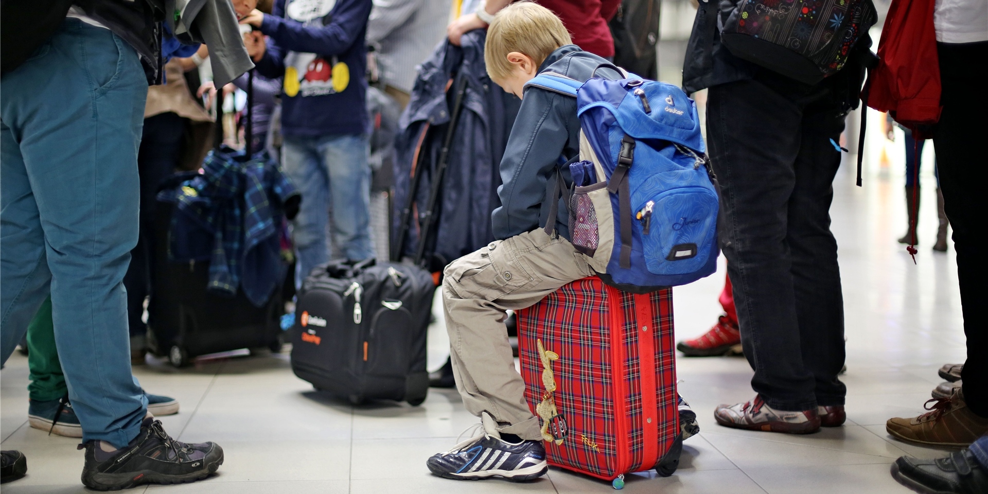 Ein kleiner Passagier sitzt auf seinem Koffer in der Wartschlange vor der Sicherheitskontrolle am Flughafen.