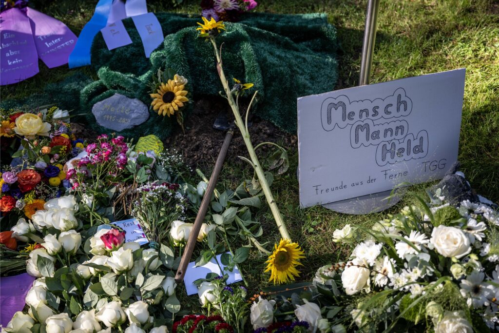 Ein Plakat mit der Aufschrift „Mensch, Mann, Held“ liegt neben Blumen und Kränzen am Urnengrab des verstorbenen Malte C. am Waldfriedhof Lauheide.