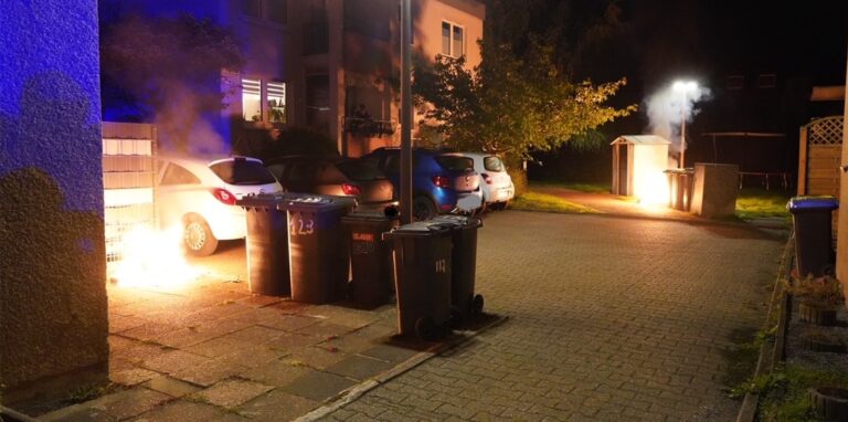 In Castrop-Rauxel, Sonnenschein, brennen zwei Mülltonnen. Dazwischen parken Autos.