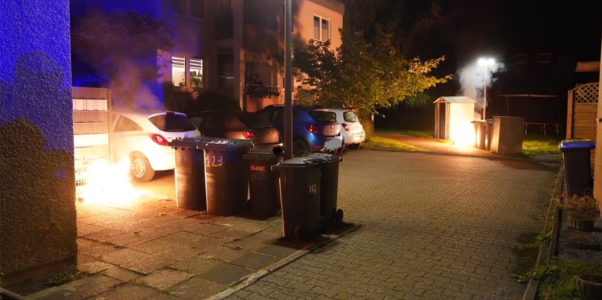 In Castrop-Rauxel, Sonnenschein, brennen zwei Mülltonnen. Dazwischen parken Autos.