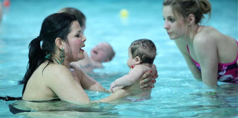 Eine Mutter hält ihren Säugling fest und lässt es vorsichtig durch das Wasser eines Schwimmbeckens gleiten.