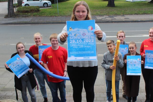 Anja Vonnahme und hinter ihr das Team von DLRG und KiJuPa Waltrop halten Plakate in die Höhe, die aufs Sponsorenschwimmen hinweisen.