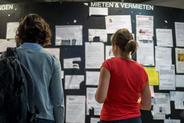 Zwei Studentinnen betrachten die Wohnungsanzeigen am Schwarzen Brett einer Universität.