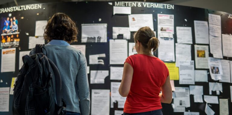 Zwei Studentinnen betrachten die Wohnungsanzeigen am Schwarzen Brett einer Universität.