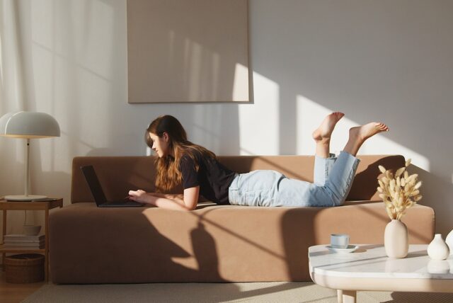 Eine Frau liegt auf einem Sofa und tippt auf ihrem Laptop, während an der Wand ein Bild hängt.