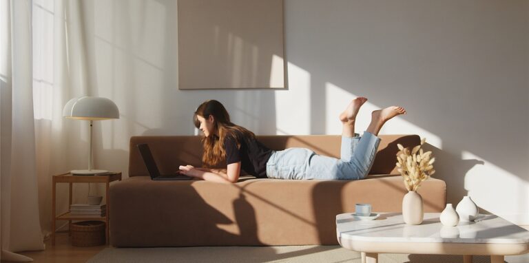 Eine Frau liegt auf einem Sofa und tippt auf ihrem Laptop, während an der Wand ein Bild hängt.