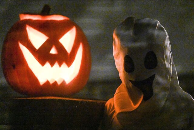 Ein Kürbis steht zu Halloween neben einem als Geist kostümierten Kind vor einem Haus.