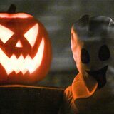 Ein Kürbis steht zu Halloween neben einem als Geist kostümierten Kind vor einem Haus.