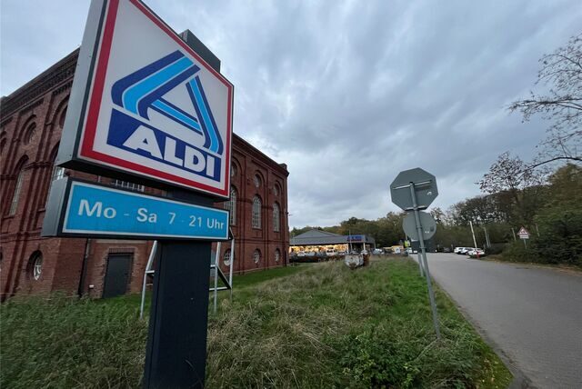 Vor einem ehemaligen Zechenhaus aus rotem Backstein steht ein Schild mit der Aufschrift „Aldi“. Auf einem kleineren Schild darunter steht „Mo – Sa 7 – 21 Uhr“. Neben Zechenhaus und einem Grünstreifen führt eine Einfahrt zu einem Aldi-Markt im Hintergrund.