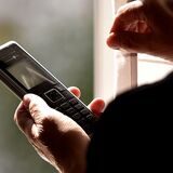 Eine Frau mit Telefon in der Hand steht vor einem Fenster. Auf der anderen Seite erkennt man schemenhaft einen telefonierenden Mann.