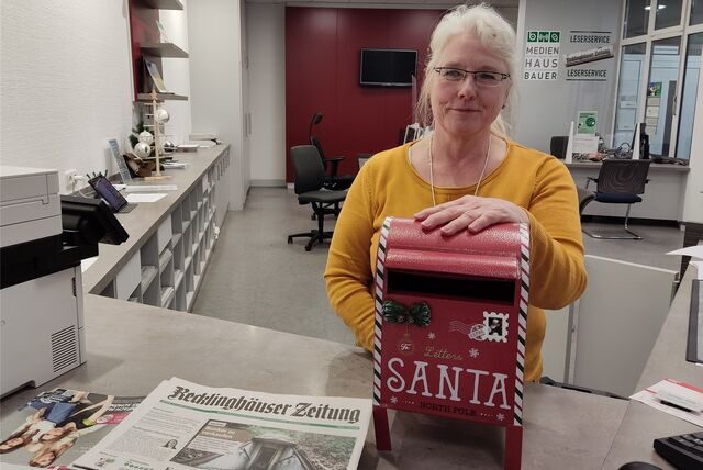 Eine Frau im gelben Pullover legt ihre Hand auf einen kleinen roten Briefkasten mit der Aufschrift „Santa“.