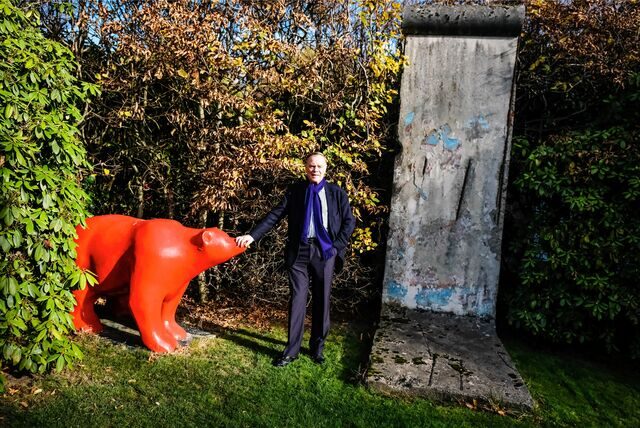 Ein Mann in Anzug legt vor einer Hecke in Recklinghausen einer roten Bärenfigur seine Hand auf die Nase, auf seiner anderen Seite steht ein Stück der Berliner Mauer.