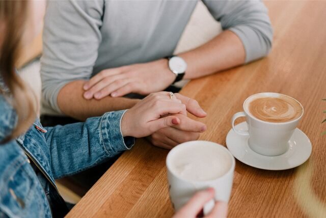 Zwei Menschen sitzen im Café und halten sich an den Händen, vor ihnen stehen zwei Tassen Cappuccino