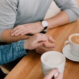 Zwei Menschen sitzen im Café und halten sich an den Händen, vor ihnen stehen zwei Tassen Cappuccino