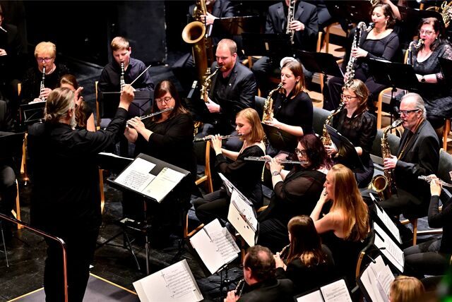 Musikerinnen und Musiker mit Blasinstrumenten auf einem Konzertpodium, davor ein Dirigent.