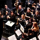 Musikerinnen und Musiker mit Blasinstrumenten auf einem Konzertpodium, davor ein Dirigent.
