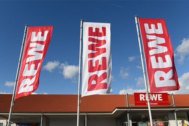 Fahnen mit dem Logo des Lebensmitteleinzelhändlers Rewe wehen vor einer Filiale.