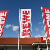 Fahnen mit dem Logo des Lebensmitteleinzelhändlers Rewe wehen vor einer Filiale.
