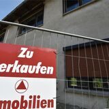 An einem Bauzaun vor einem Haus hängt ein Schild mit der Aufschrift „zu verkaufen“.