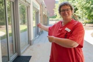 Eine Frau steht auf einer Terrasse und zeigt auf die Fenster des Hauses.