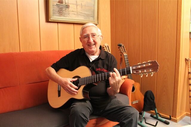 Der 91-jährige Norbert Thamm sitzt in seinem Wohnzimmer auf einem Sofa und hält eine Gitarre in der Hand.