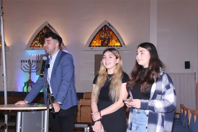 Ein junger Mann steht in einer Synagoge an einem Rednerpult, neben ihm zwei junge Frauen.