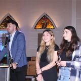 Ein junger Mann steht in einer Synagoge an einem Rednerpult, neben ihm zwei junge Frauen.