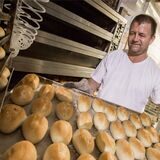 Bäcker vor einem Ofen hält ein Blech mit Brötchen-Teiglingen.