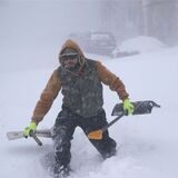 Ein Mann stapft mit zwei Schaufeln über eine Schneewehe, um einem gestrandeten Autofahrer zu helfen.