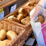 Eine Bäckerei-Verkäuferin nimmt mit einem Handschuh ein Brötchen aus einem Korb und legt es in eine Papiertüte.