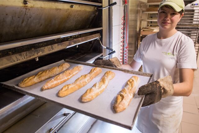 Bäckerin vor einem Ofen hält ein Blech mit Baguettes.
