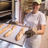 Bäckerin vor einem Ofen hält ein Blech mit Baguettes.