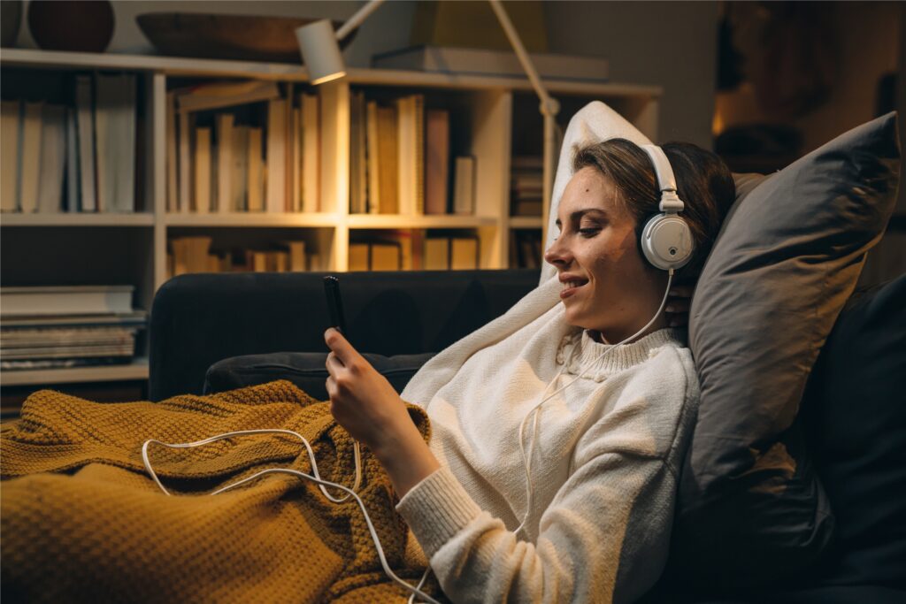 frau liegt gemütlich unter einer decke auf der couch und hat kopfhörer auf frau liegt gemütlich unter einer decke auf der couch und hat kopfhörer auf