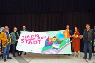 Die Organisatoren des Familienfestes halten ein großes Transparent mit dem Schriftzug „Der Zug kommt in die Stadt“ in den Händen.