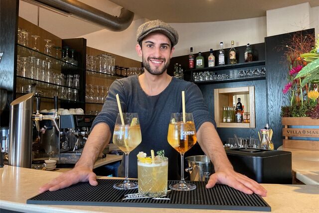 Ein Mann mit Schiebermütze steht hinter einer Theke in einer Gastronomie in Recklinghausen. Vor ihm auf der Theke stehen drei Gläser mit verschiedenen alkoholischen Mix-Getränken.