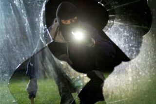 Ein maskierter Mann mit Taschenlampe leuchtet durch eine zerbrochene Scheibe.