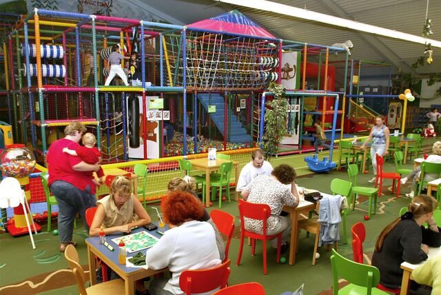 Eltern sitzen an Tischen in einem Indoor-Spielplatz, Kinder spielen im Hintergund.
