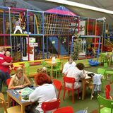 Eltern sitzen an Tischen in einem Indoor-Spielplatz, Kinder spielen im Hintergund.